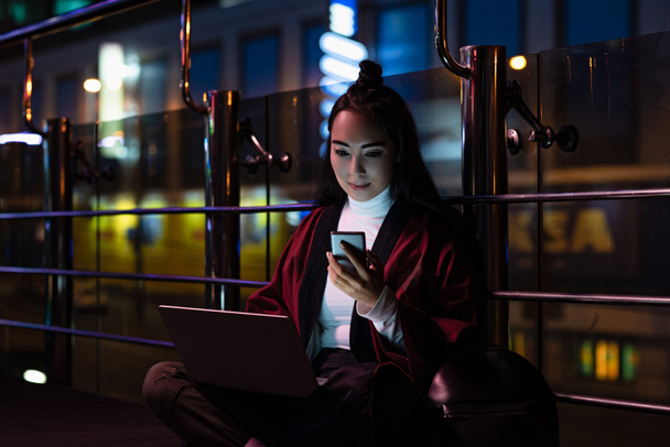 gyönyörű ázsiai lány ül, és a smartphone utcán neon fénnyel, város a jövőbeni koncepció laptop segítségével kimonó - Fotó, kép