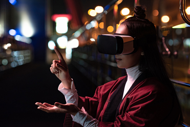 привлекательная азиатская девушка, использующая гарнитуру виртуальной реальности и указывающая на что-то на улице вечером, город будущей концепции
 - Фото, изображение