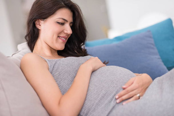young happy pregnant woman keeping hand on belly while sitting on sofa at home - Photo, Image
