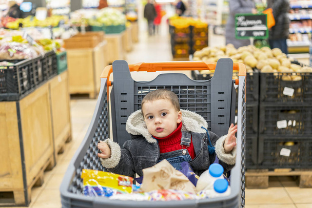 Boy 2 years in a supermarket, sitting in a cart full of various products. - Foto, Bild