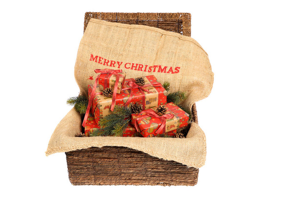 Christmas wrapped gifts with red ribbons on a jute sack with text "Merry Christmas", decorated with pine cones and pine twigs in an open wicker basket, isolated on white background  - Photo, Image