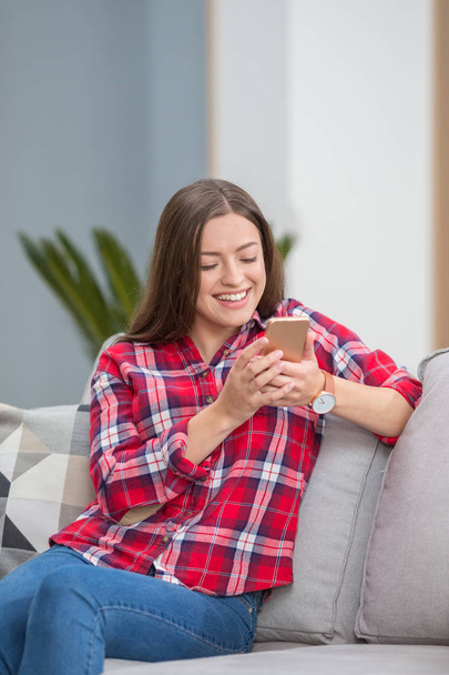 Good-looking, magnificent, exquisite, delicate, winsome, lovable woman sit on cozy couch in shirt and denim jeans in modern light interior and look at phone screen make hollywood smile - 写真・画像