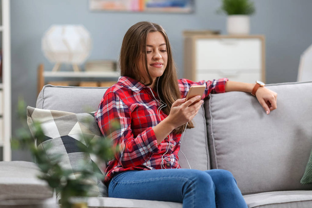Female sits on sofa and looks at phone. She is listening music through headphones. Girl holds one of her hands on headphone. She smiles. - Photo, Image