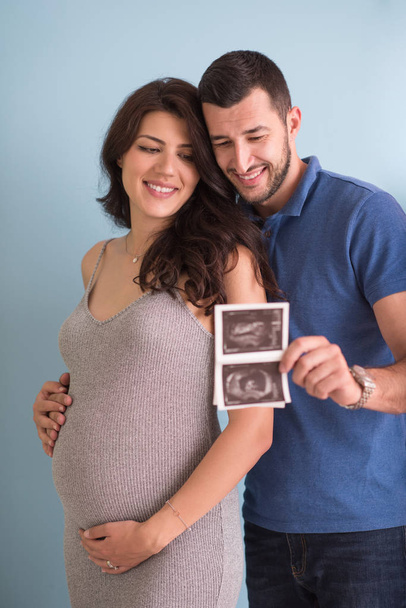 happy pregnant couple looking ultrasound picture of their unborn baby, isolated on blue background - 写真・画像