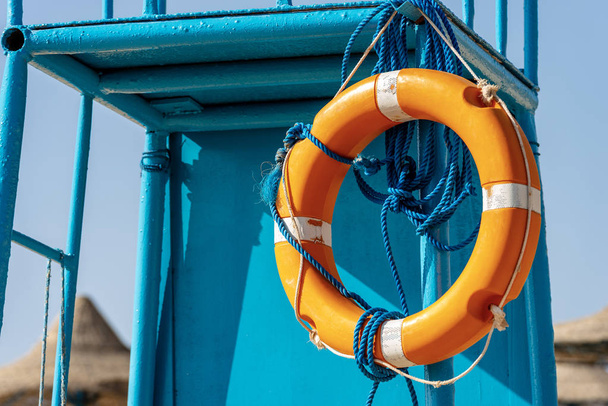 Close-up of an orange and white lifebuoy with ropes hanging on a blue lifeguard chair - Photo, Image