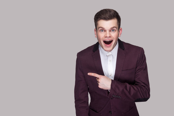 Portrait of surprised handsome young man in violet suit and white shirt looking at camera and pointing aside with amazed face on grey background - Foto, Imagen