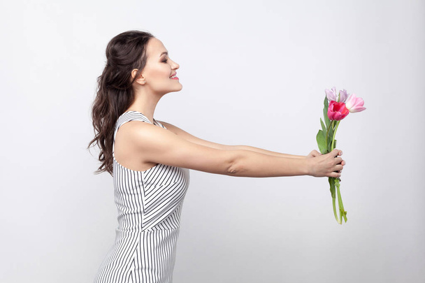 perfil vista lateral retrato de joven hermosa mujer en vestido blanco a rayas de pie y dando tulipanes sobre fondo gris
. - Foto, Imagen