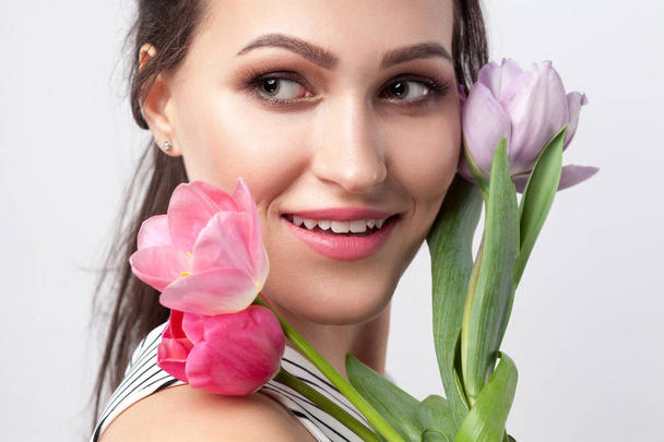 portrait of young brunette beautiful woman with makeup in striped dress holding tulips and looking away with toothy smile on grey background. - Foto, Imagem