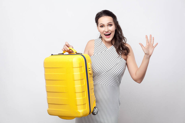 Portrait of happy beautiful young traveller in striped dress holding yellow suitcase and ready for vacation on grey background - Fotografie, Obrázek