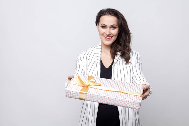 Kindness attractive young brunette woman in white coat standing and giving present with yellow bow while looking at camera on grey background - Foto, imagen