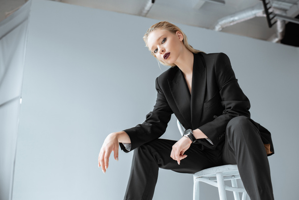 beautiful blonde young woman in trendy black suit sitting on chair on grey - Foto, Imagen