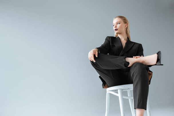 attractive fashionable girl in black suit sitting on chair isolated on grey - Photo, Image