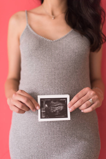 smiling pregnant woman showing ultrasound picture of her unborn baby isolated on red background - Photo, image