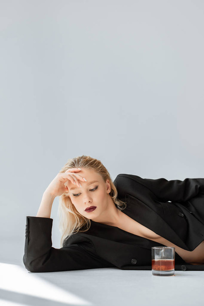 modelo bonito em terno moderno preto deitado no cinza com vidro de uísque
 - Foto, Imagem