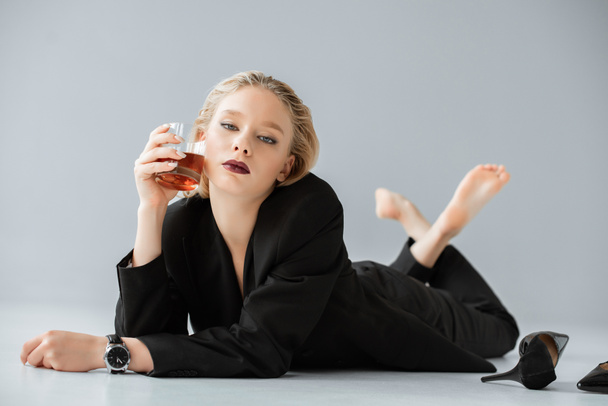 beautiful fashionable woman in black trendy suit holding glass of whiskey and lying on grey - Fotó, kép