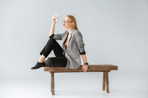 stylish girl holding glass of milk and sitting on wooden bench on grey - Фото, зображення