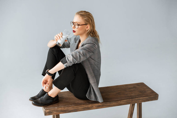beautiful fashionable woman drinking milk from glass while sitting on wooden bench isolated on grey - 写真・画像