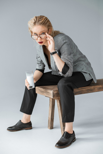chica de moda en gafas con vaso de leche y sentado en un banco de madera en gris
 - Foto, Imagen