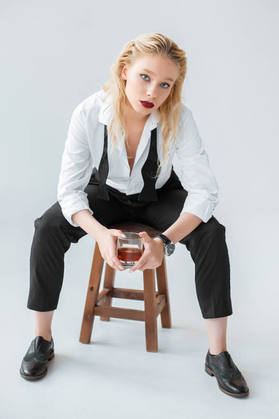 ελκυστικό μοντέλο μόδας με ποτήρι ουίσκι που κάθεται στο σκαμνί σε γκρι - Φωτογραφία, εικόνα