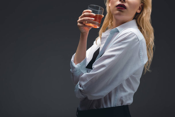 bijgesneden weergave van meisje in formele slijtage holding glas cognac, geïsoleerd op donkergrijs - Foto, afbeelding