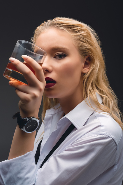 όμορφο κομψό κορίτσι σε επίσημη ένδυση κρατώντας το ποτήρι του ουίσκι, απομονώνονται σε σκούρο γκρι - Φωτογραφία, εικόνα