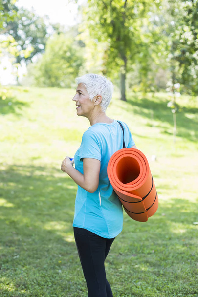 Portrait of senior woman holds fitness mat on her back in the park and preparing for exercise - Photo, Image