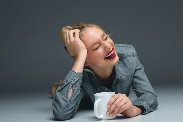 beautiful laughing girl with cup of coffee lying on grey - Photo, Image