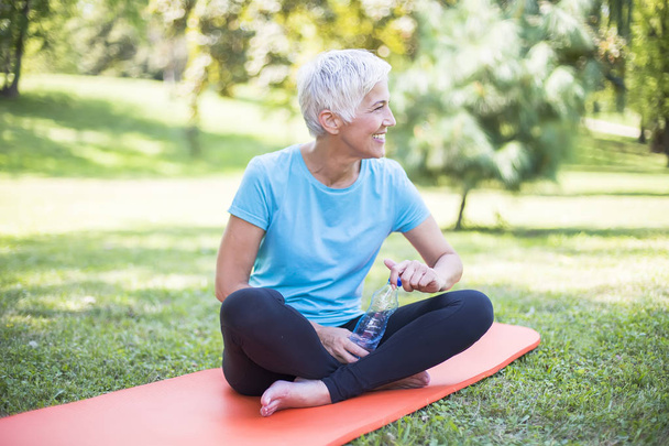 Senior sportive woman sitting on mat outside and resting after workout - Photo, Image