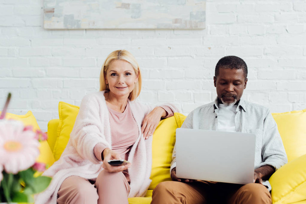 mature african american man using laptop while woman sitting on sofa with remote controller - Foto, Bild