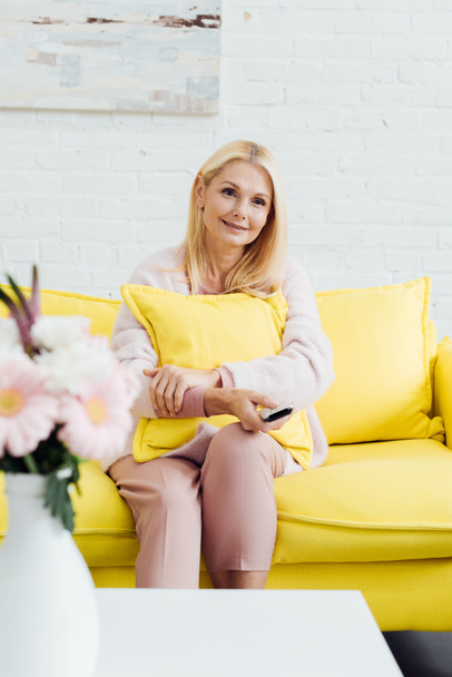 beautiful smiling mature woman sitting on bright yellow sofa with tv remote controller - Foto, Imagen