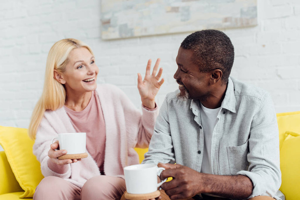 emotional mature woman sitting on sofa and talking with african american man with white cups - Foto, immagini