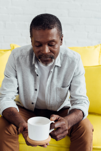 handsome african american man sitting on bright yellow sofa with white cup - Foto, imagen