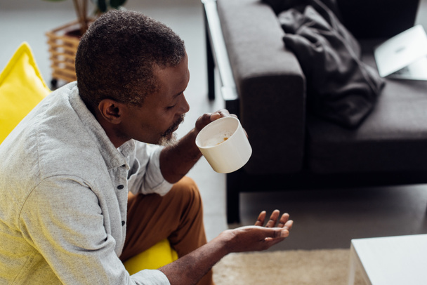 mature african american man sitting on sofa and drinking coffee in living room  - Photo, Image