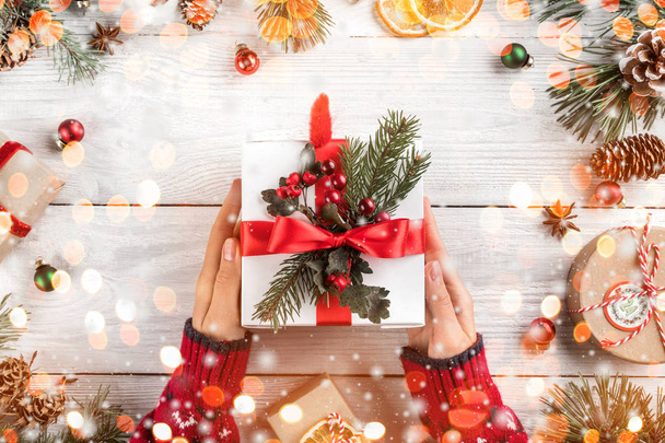 weibliche Hände halten Weihnachtsgeschenkschachtel auf weißem Holzgrund mit Tannenzweigen, Tannenzapfen. Weihnachten und ein frohes neues Jahr Thema, Bokeh, funkeln, glühen. flache Lage, Draufsicht - Foto, Bild
