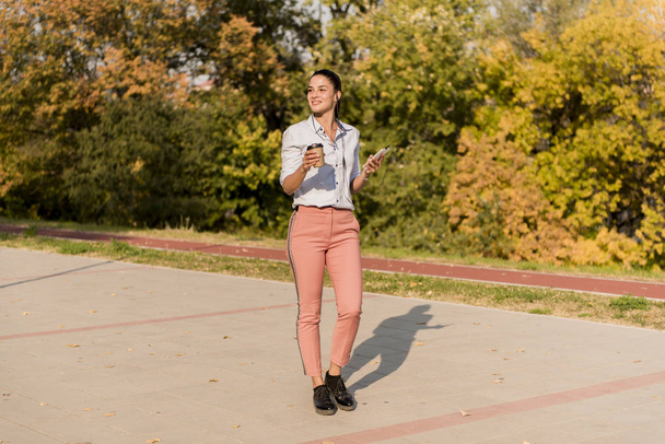 Boldog fiatal nő kávé hallgat zenét a smartphone és a séta a sétány ta gyönyörű őszi nap - Fotó, kép