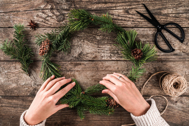 Woman hands making a Christmas wreath of fir branches and pine cones on wooden background with scissors and skein of jute. Xmas and Happy New Year card. Flat lay, top view - Fotoğraf, Görsel