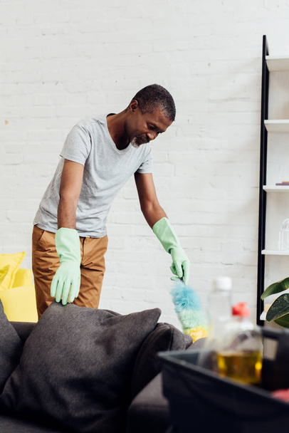 красивий афроамериканський чоловік в гумових рукавичках робить прибирання будинку з пилом
 - Фото, зображення