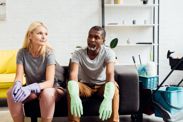 tired couple in rubber gloves sitting on sofa after housecleaning - Photo, Image