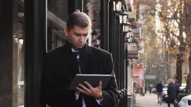Młody człowiek w garniturze i czarny płaszcz wykorzystuje cyfrowy tablicowy - Materiał filmowy, wideo