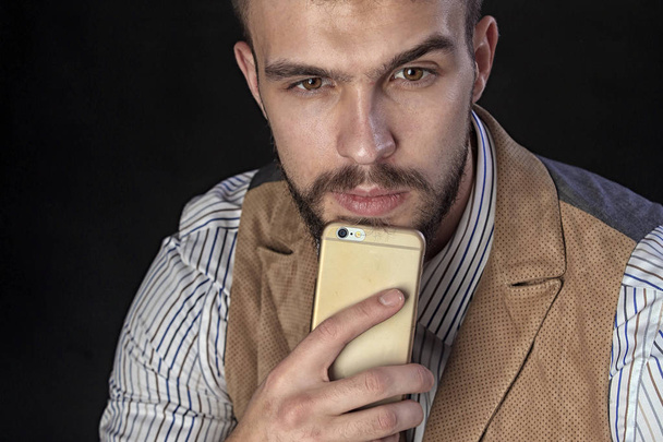 closeup portrait of the beautiful charismatic young man with a beard, with phone wearing shirt, vest and tie on a dark background studio - Foto, Bild