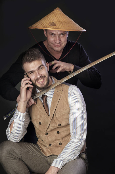 portrait of a man in the Vietnamese hat sword threatening the young man with a smartphone on dark background studio - Foto, immagini
