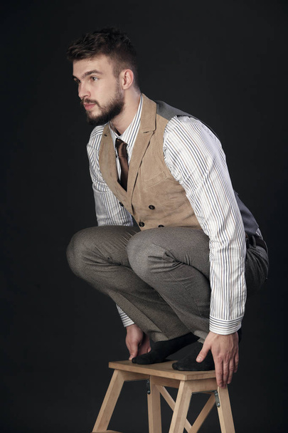 closeup portrait of the beautiful charismatic young man with a beard, squatted on a stool on a dark background studio  - Foto, Bild