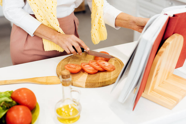 close up of woman slicing tomatoes and reading recipe in cookbook at kitchen - Photo, Image