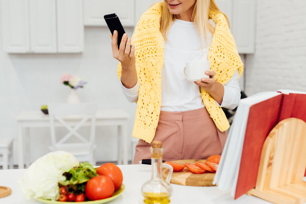 cropped view of blonde woman holding smartphone while cooking dinner - Fotoğraf, Görsel