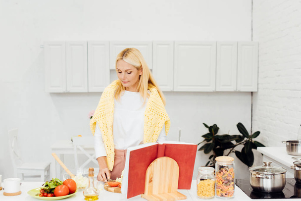 beautiful blonde woman cooking salad with cookbook at kitchen - Photo, Image