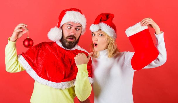 Christmas stocking tradition. Christmas celebration concept. Woman and bearded man in santa hat waiting christmas red background. Couple christmas santa costume hold sock and ornament ball. - Фото, зображення