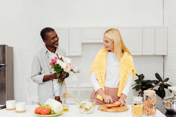 キッチンで成熟した金髪妻に花を gifting ハンサムなアフリカ系アメリカ人の夫 - 写真・画像