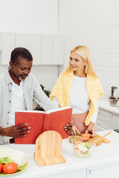 happy interracial couple cooking dinner together and reading recipe in cookbook at kitchen - Φωτογραφία, εικόνα