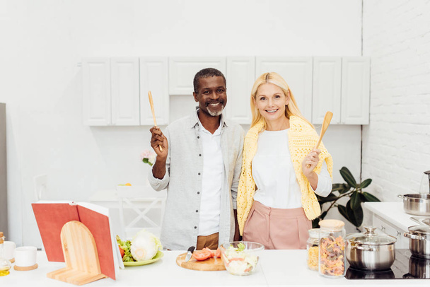 cheerful interracial couple holding cooking utensils together at kitchen - Fotoğraf, Görsel