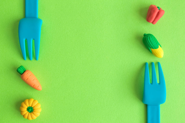 Flat lay of plastic forks with small rubber vegetable toys against green background minimal creative vegetarian concept. - Foto, imagen